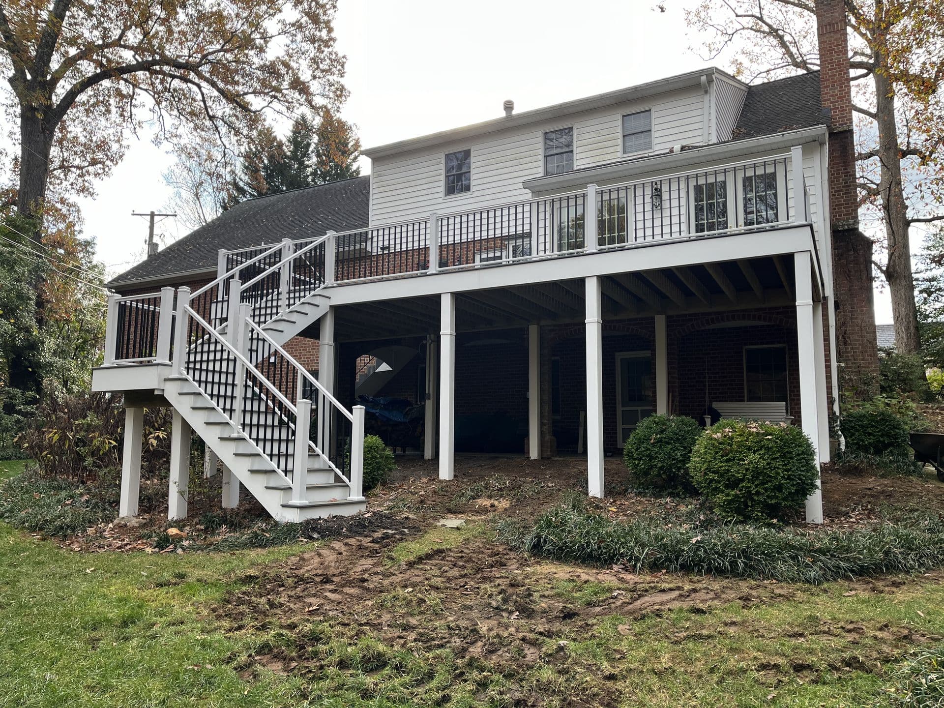 Two-story house with elevated deck and white railings, surrounded by autumn trees and greenery. Stairs lead down to a grassy backyard, conveying tranquility.