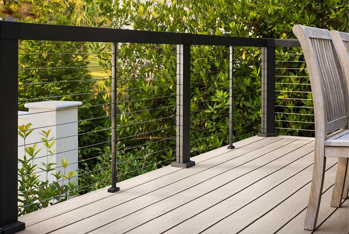 A peaceful deck with light wooden flooring features a sleek black metal railing. Lush green leaves create a serene, natural backdrop on a sunny day.