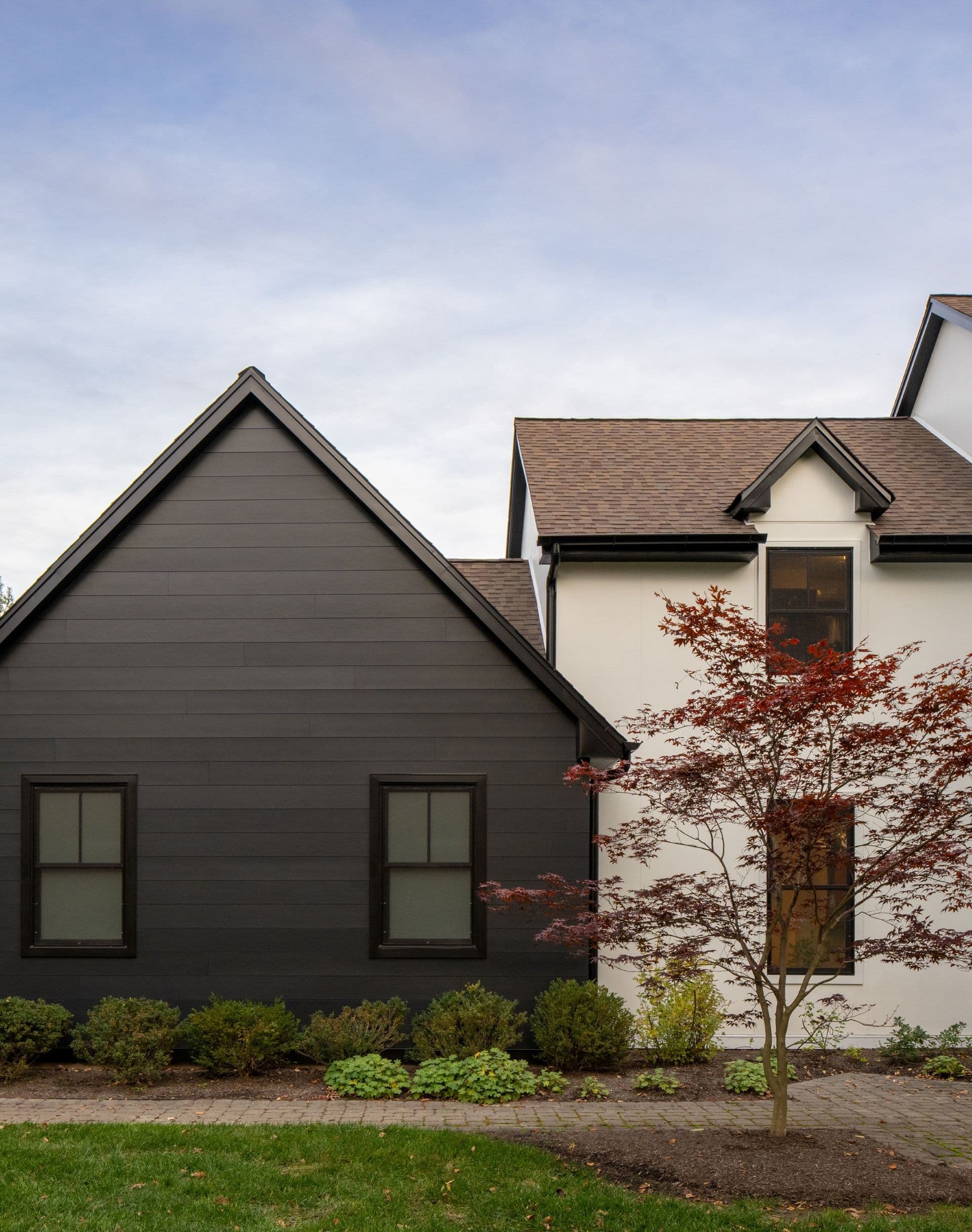 Modern house exterior with a contrasting design: one section is painted black with dark windows, the other white with brown roofing. A small red-leafed tree stands in front, conveying a minimalist yet cozy atmosphere.