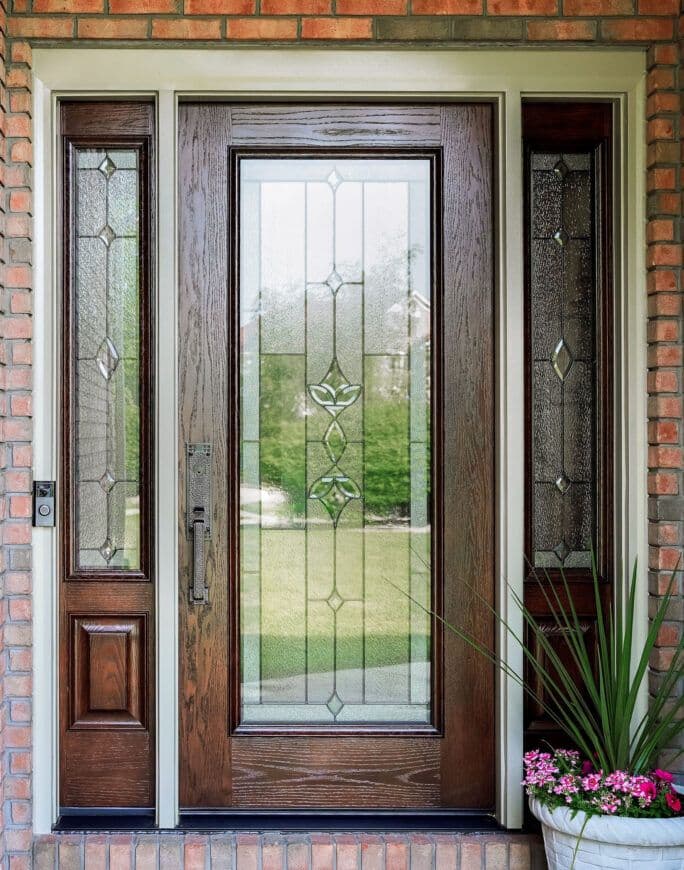 A wooden door with decorative glass panels, flanked by two side windows, set in a brick wall. A potted plant with flowers sits to the right.
