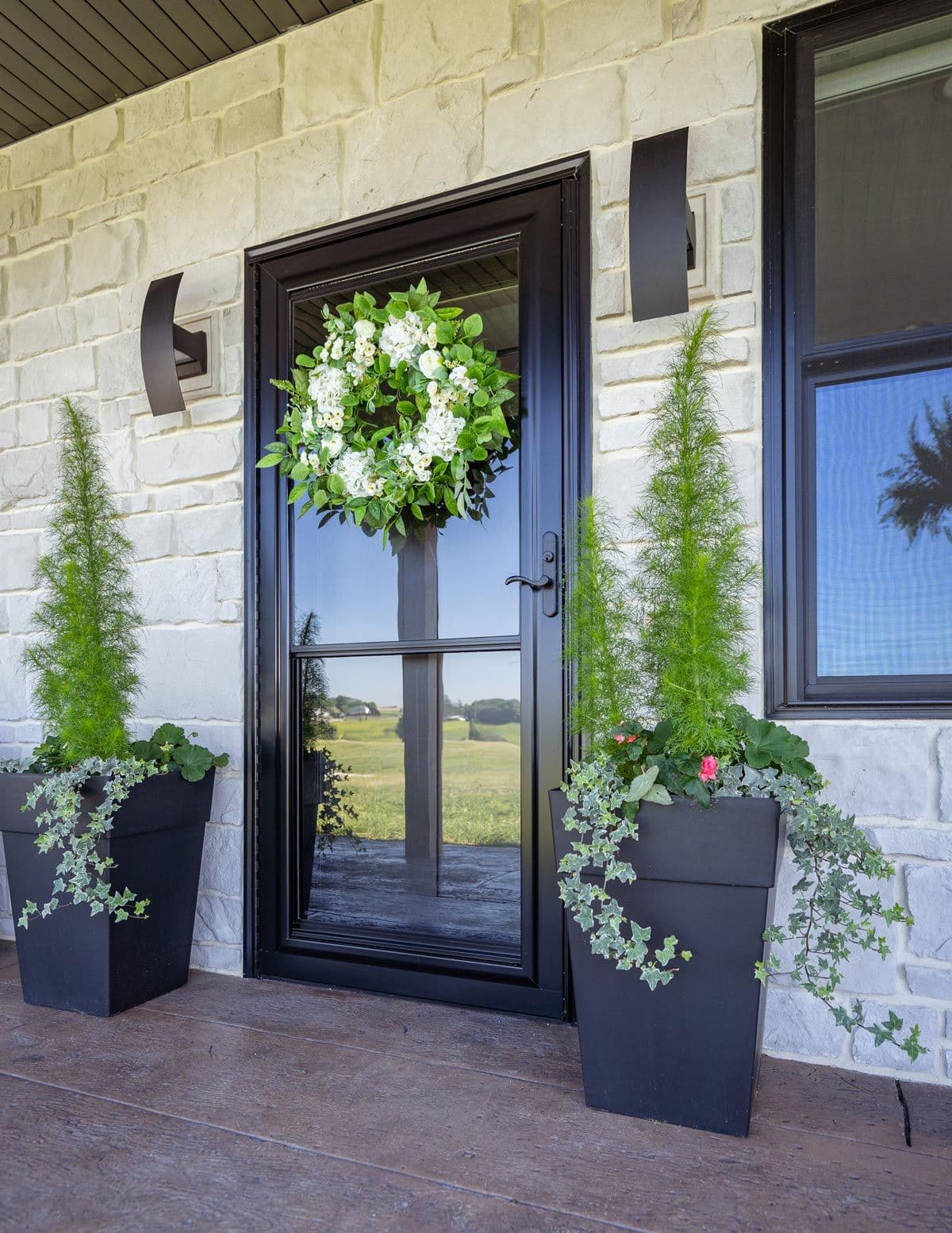 Black front door with glass panels, adorned with a vibrant green and white wreath. Flanked by two tall potted plants, set against a stone wall.