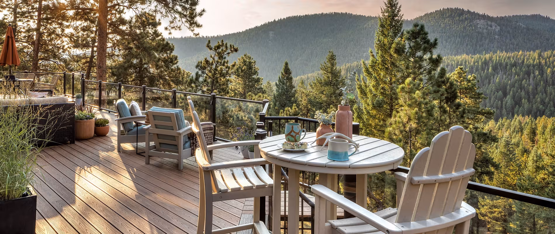 A wooden deck overlooking a lush forest and mountains. White chairs surround a table with cups and a thermos, evoking a peaceful, cozy retreat vibe.