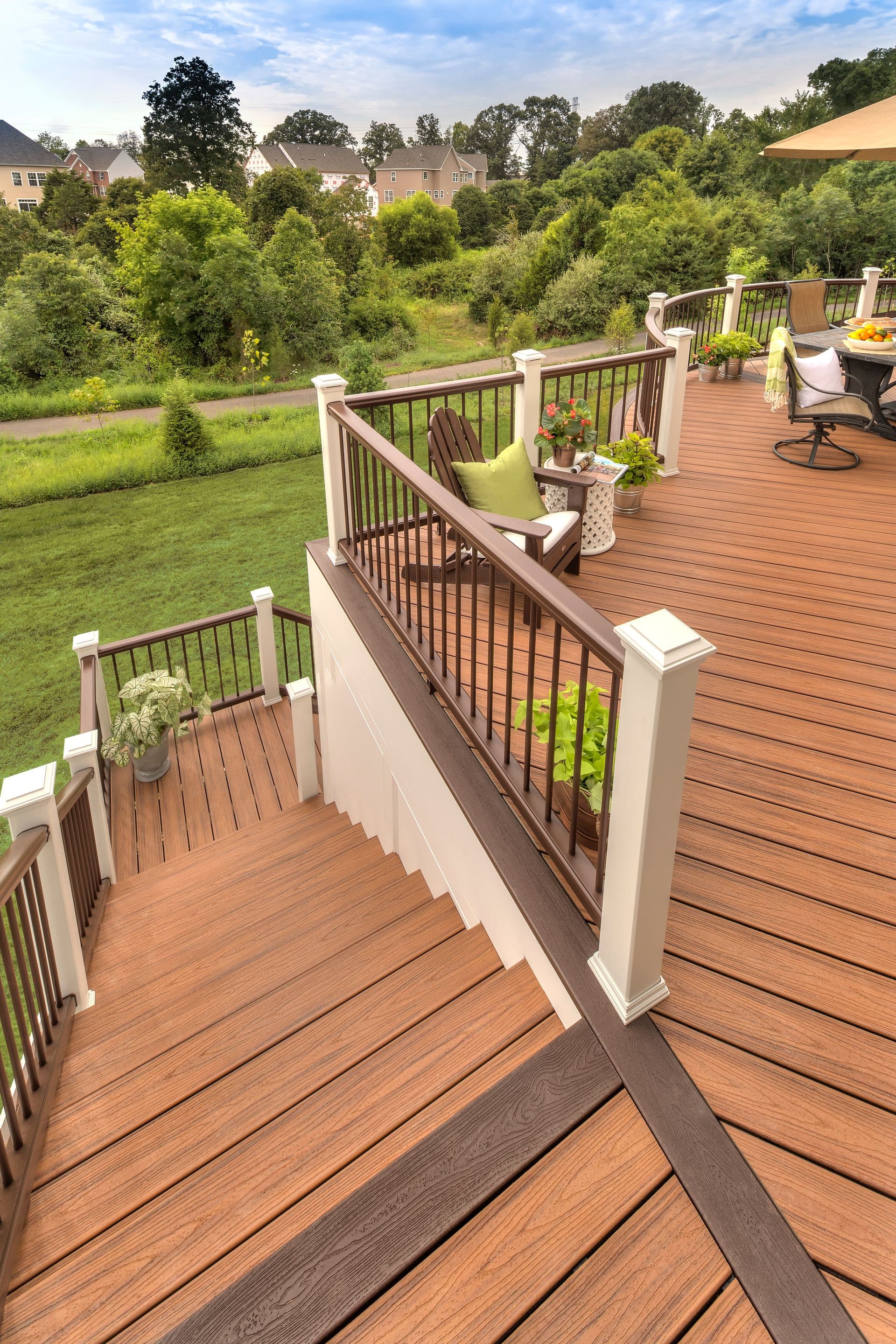 Elevated wooden deck with railing overlooks a lush green backyard and trees. The deck has chairs, plants, and a table, creating a relaxing outdoor space.
