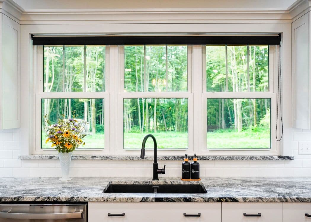 Bright kitchen with a large window overlooking green trees. A granite countertop holds a vase of flowers and two soap bottles. A black faucet adds contrast.