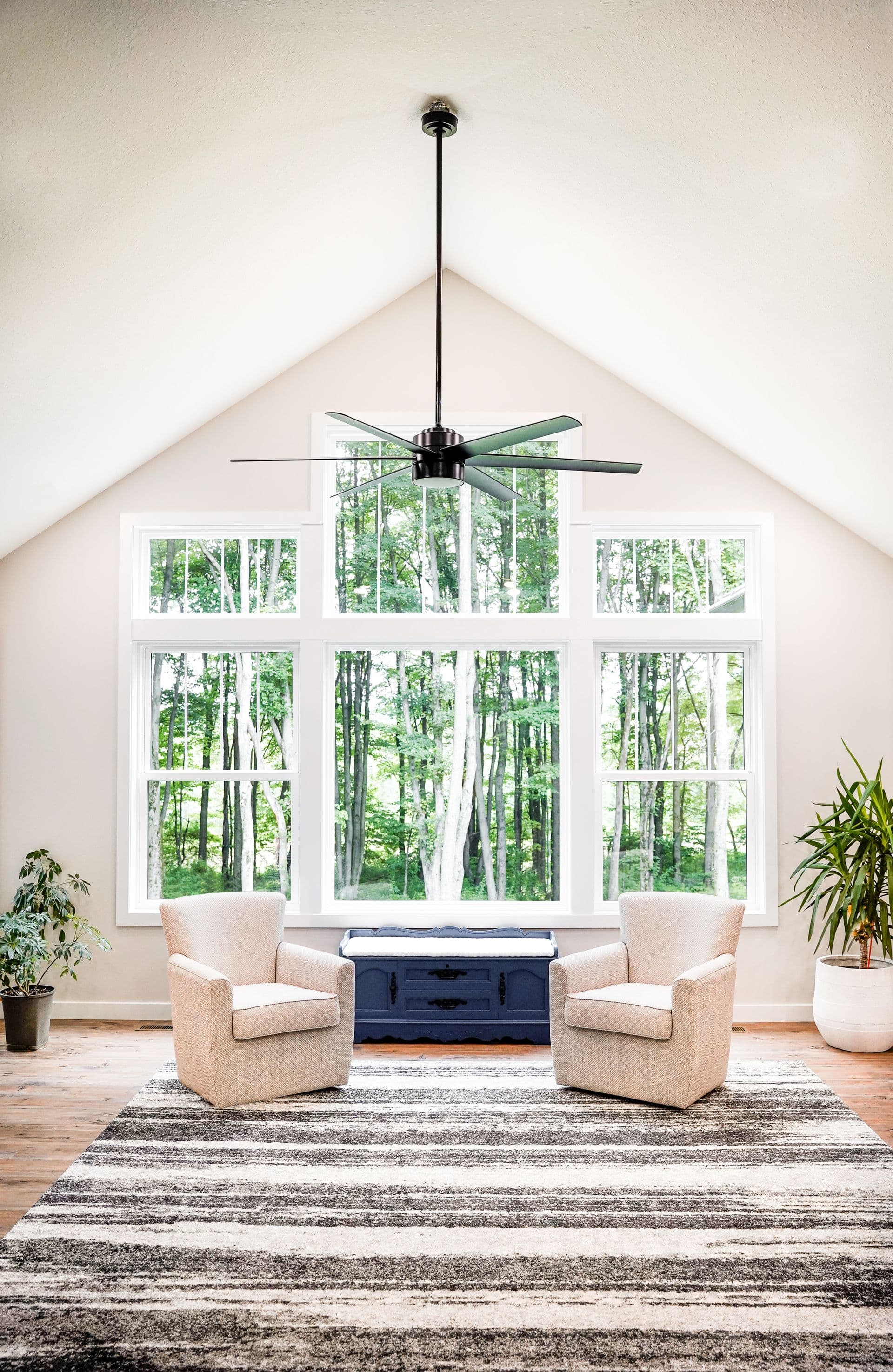 Bright living room with two beige armchairs facing large windows with forest view. A ceiling fan hangs above, and green plants accent the space.